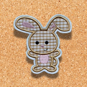 May include: An embroidered bunny patch with a light blue border. The bunny has a brown and white checkered body, pink accents, and black eyes. The patch is on a corkboard background.