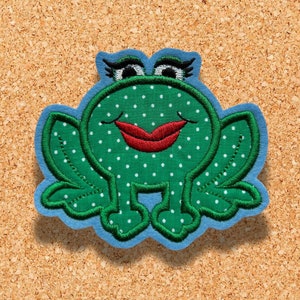 Op de afbeelding: Groene vilt applicatie van een kikker met witte stippen, rode lippen en blauwe wimpers. De kikker zit op een kurk achtergrond.