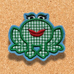 Könnte beinhalten: Grüner und weißer Gingham-Frosch mit roten Lippen und blauem Umriss. Der Frosch sitzt auf einem Korkuntergrund.