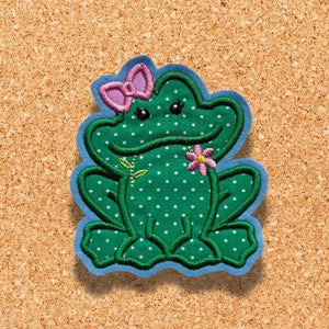 Op de afbeelding: Groene kikker met witte stippen, met een roze strik en een roze bloem. De kikker zit op een blauwe achtergrond.