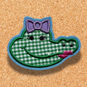 Puede incluir: Cabeza de cocodrilo de tela vichy verde y blanca con un lazo morado, bordada con ojos negros y una boca rosa.