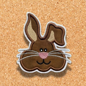 May include: Brown bunny rabbit face with white outline and pink nose. The bunny has two black eyes and a small smile.