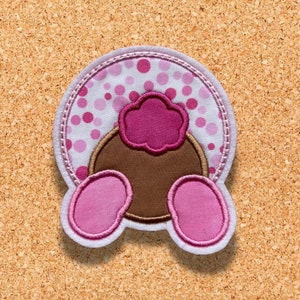 May include: A pink and brown felt applique of a bunny's rear end with a pink flower on top. The bunny's tail is white with pink polka dots.