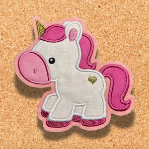 Könnte beinhalten: Ein rosa-weißes Einhorn-Applikationsmotiv mit goldenem Horn und einem kleinen goldenen Herz an der Seite. Das Einhorn hat eine rosa Mähne und einen rosa Schwanz.