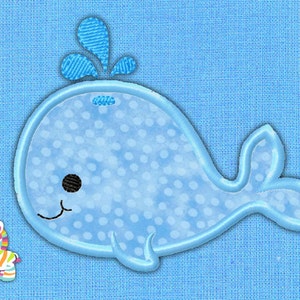 Puede incluir: Una ballena azul con lunares blancos y un chorro de agua en la parte superior, bordada sobre un fondo de tela azul. Un pequeño juguete de cebra está en la esquina inferior izquierda.
