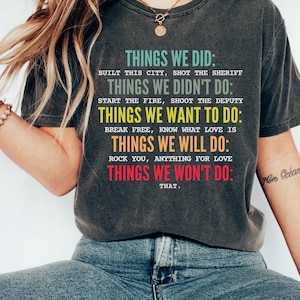 May include: Dark gray t-shirt with the text "THINGS WE DID: BUILT THIS CITY, SHOT THE SHERIFF" and other phrases in a colorful font. The shirt is worn with blue jeans. The overall theme is a casual, statement tee.