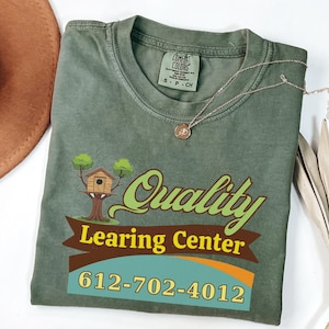 Könnte beinhalten: Olivgrünes T-Shirt mit Grafik für ein Lernzentrum. Das Design umfasst ein Baumhaus, die Worte "Quality Learning Center" und eine Telefonnummer. Eine goldene Halskette und ein brauner Hut sind ebenfalls im Bild.