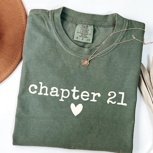 Peut inclure: Un t-shirt vert olive avec le texte "chapter 21" et un cœur blanc. Un collier en or avec un pendentif repose sur le t-shirt. Un chapeau marron et des feuilles de palmier séchées sont en arrière-plan.