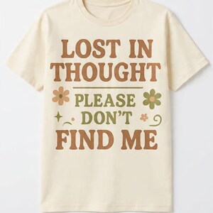 May include: Cream-colored t-shirt with the text "LOST IN THOUGHT PLEASE DON'T FIND ME" in brown and green. The design includes flower and star graphics. A casual, short-sleeved tee.