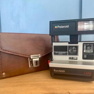 May include: A vintage Polaroid Sun 600 LMS instant camera with a black and silver body, and a brown leather carrying case. The camera has a built-in flash and the Polaroid logo. The case has a metal clasp.