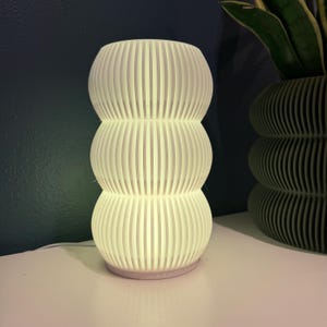 May include: A white, stacked, ribbed table lamp with a soft glow. The lamp has a rounded base and three stacked sections. The lamp is on a white surface with a dark blue background. A matching ribbed planter is visible in the background.
