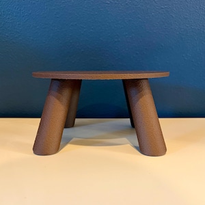 Pode incluir: Pequena mesa de madeira castanha com tampo redondo e quatro pernas anguladas. A mesa é de cor castanho escuro e tem um design simples e minimalista. Adequada para exibir pequenos objetos.