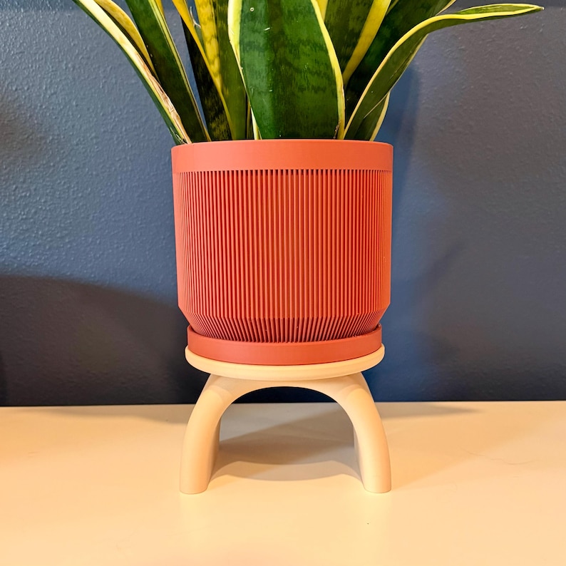 May include: A cylindrical terracotta-colored planter with a vertical ribbed texture, resting on a light beige arched stand. A green and yellow snake plant grows out of the top of the planter, which features a distinct horizontal rim.