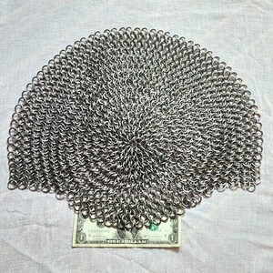 May include: A circular piece of silver-colored chainmail constructed from numerous interlocking metal rings. The chainmail is positioned directly above a single United States one-dollar bill, which serves as a visual reference for its scale.
