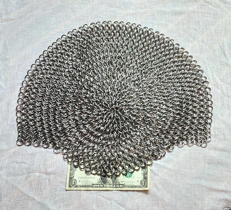 May include: A circular piece of silver-colored chainmail constructed from numerous interlocking metal rings. The chainmail is positioned directly above a single United States one-dollar bill, which serves as a visual reference for its scale.
