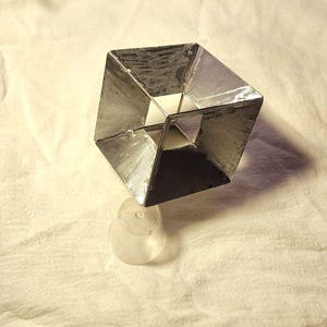 May include: A geometric sculpture featuring a silver-toned, open-frame cube structure with metallic panels, supported by a clear, circular plastic base. The object displays a reflective surface and sharp, angular lines, creating a hollow, three-dimensional form.