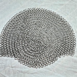 May include: A circular piece of chainmail armor constructed from numerous small, interlocking silver-colored metal rings. The pattern radiates outward from the center, creating a dense, uniform mesh structure with a metallic, reflective finish.