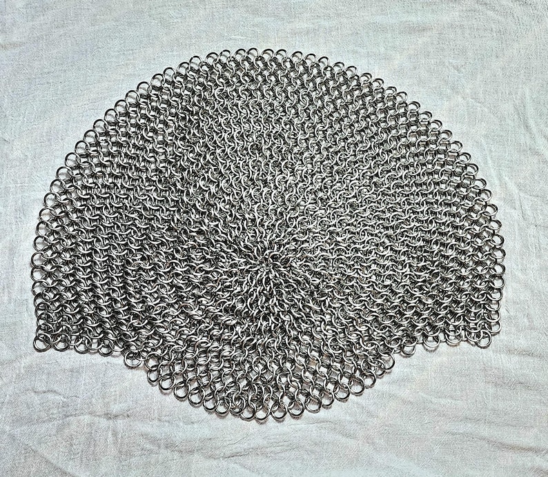 May include: A circular piece of chainmail armor constructed from numerous small, interlocking silver-colored metal rings. The pattern radiates outward from the center, creating a dense, uniform mesh structure with a metallic, reflective finish.