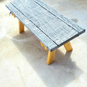 May include: A rectangular wooden bench featuring a top surface with a distressed black and white finish. The bench is supported by four sturdy, natural light-brown wooden legs that are angled outward to provide stability for the seat structure.