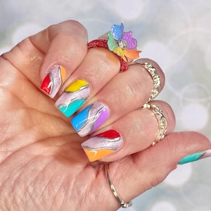 Peut inclure: Des ongles de forme carrée arborant un motif multicolore avec des lignes ondulées à paillettes argentées séparant des blocs de vernis rouge, orange, jaune, turquoise, bleu et violet. Une bague rouge avec une fleur multicolore est portée.