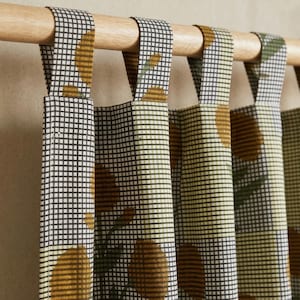 Peut inclure: Un panneau de rideau avec un motif à carreaux noirs et blancs, agrémenté de motifs floraux jaune moutarde et de feuilles vertes. Le tissu est suspendu à une tringle en bois clair par quatre pattes en tissu fixées au bord supérieur du rideau.