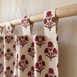May include: A cream-coloured curtain featuring a repeating pattern of dark red floral motifs with green stems and leaves. The curtain is suspended from a light-toned wooden rod by fabric tabs, creating vertical folds along the length of the fabric.