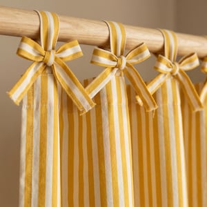 May include: Yellow and white striped curtains with matching tie-up bows. The curtains are hanging from a wooden rod. The fabric has vertical stripes of yellow and white. The bows are tied at the top of each curtain panel.