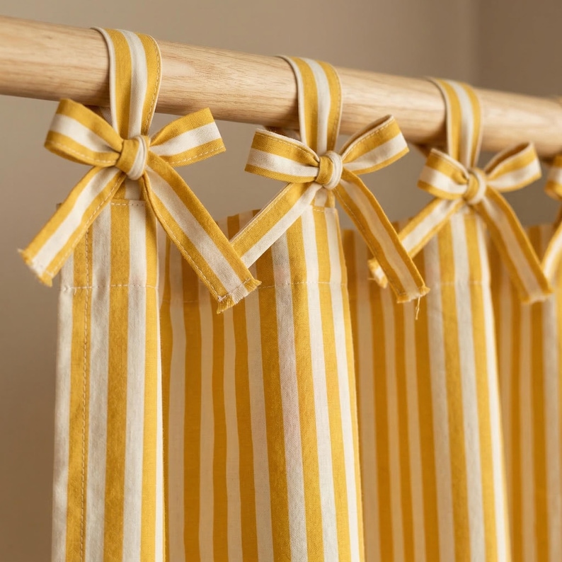 May include: Yellow and white striped curtains with matching tie-up bows. The curtains are hanging from a wooden rod. The fabric has vertical stripes of yellow and white. The bows are tied at the top of each curtain panel.