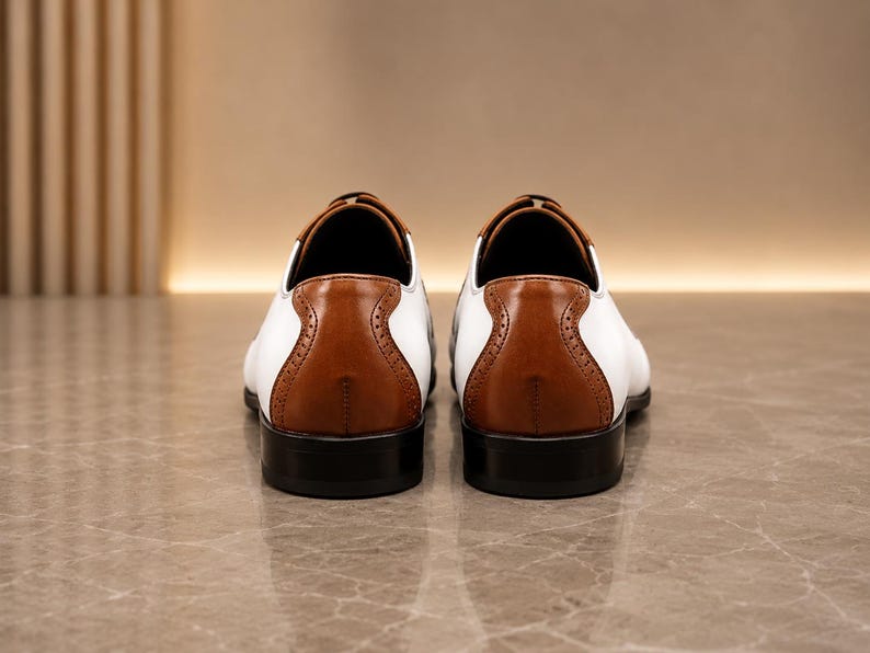 May include: A pair of white and tan leather dress shoes viewed from the back. The shoes feature a smooth white leather upper, a tan leather heel counter with decorative brogue perforations, and a solid black stacked heel and sole base.