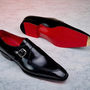 May include: A pair of black leather dress shoes with a shiny finish. The shoes feature a buckle closure and red soles. One shoe is positioned upright, while the other is lying on its side, showcasing the red sole.