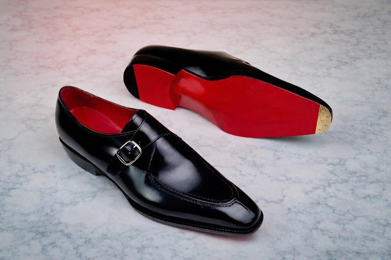 May include: A pair of black leather dress shoes with a shiny finish. The shoes feature a buckle closure and red soles. One shoe is positioned upright, while the other is lying on its side, showcasing the red sole.