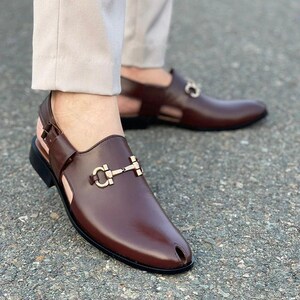 May include: Brown leather sandals with a gold-colored buckle detail. The sandals have a closed-toe design with a small opening and a strap around the ankle. The shoes are worn with light-colored pants.