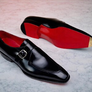 May include: A pair of black leather dress shoes with a shiny finish. The shoes feature a buckle closure and red soles. One shoe is positioned upright, while the other is lying on its side, showcasing the red sole.
