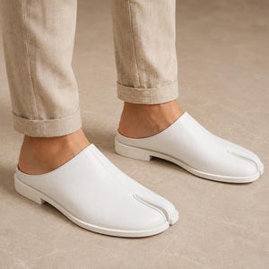 May include: A pair of white leather slip-on mules featuring a distinctive split-toe design. These flat shoes have a smooth finish, a low heel, and a seam running down the center of the vamp, providing a clean and structured appearance for footwear.