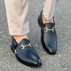 Pode incluir: Sapatos pretos de couro sem cadarços com fivela de metal dourado. Os sapatos têm bico fino e tira traseira. Os sapatos são usados com calças bege.