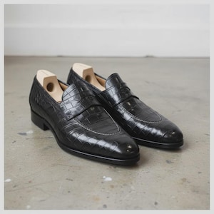 May include: A pair of black leather loafers with a crocodile texture. The shoes have a classic design with a rounded toe and a low heel. White stitching accents the edges. Shoe trees are inserted in the shoes.