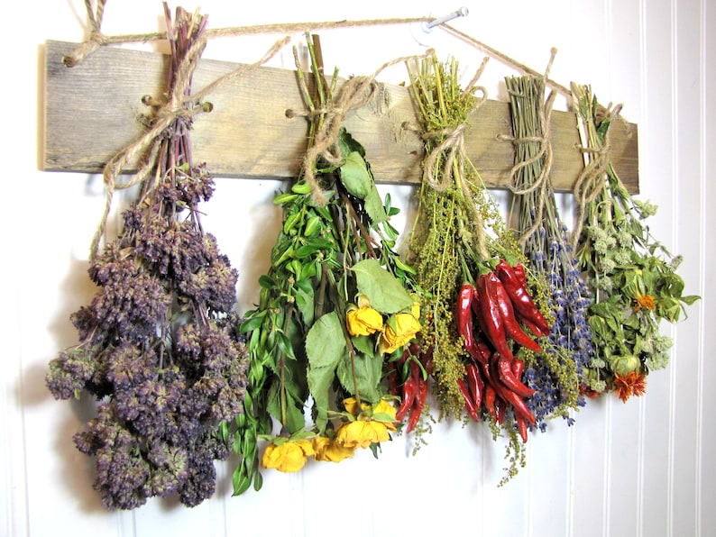 May include: A rustic wooden rack with six bundles of dried herbs and flowers hanging from it. The bundles include purple oregano, yellow roses, green herbs, red chili peppers, lavender, and dried flowers.