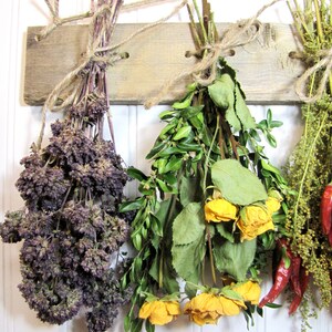 May include: Three bundles of dried herbs and flowers hanging on a rustic wooden board. The bundles are tied with twine and include purple oregano, green rosemary, and yellow roses.