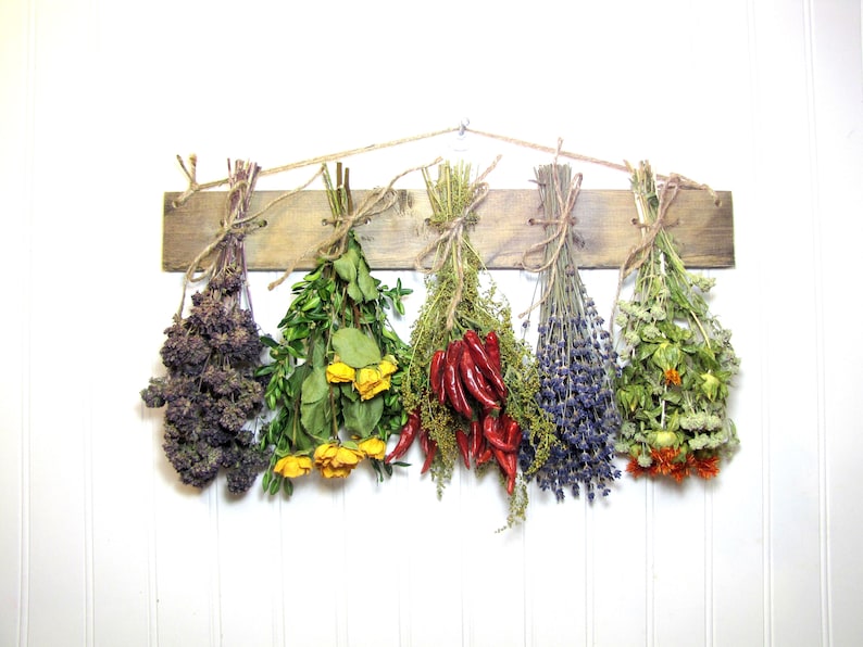 May include: Six bundles of dried herbs and peppers hanging on a wooden rack. The bundles include purple, green, yellow, red, blue, and orange dried flowers and herbs.
