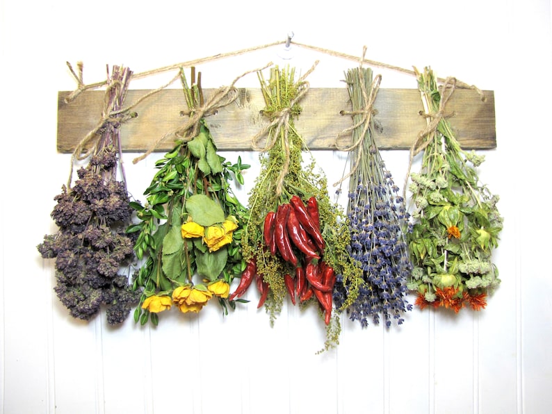 May include: Six bundles of dried herbs and flowers hanging from a wooden rack. The bundles include purple oregano, yellow roses, red chili peppers, lavender, and orange calendula.