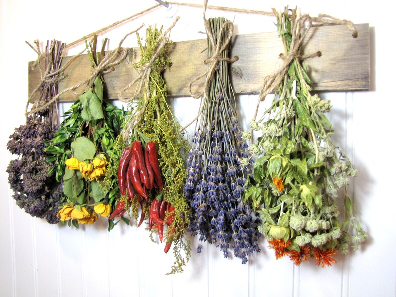 May include: A rustic wooden rack with six bundles of dried herbs and flowers hanging from it. The bundles include lavender, red chili peppers, yellow roses, and various green herbs.