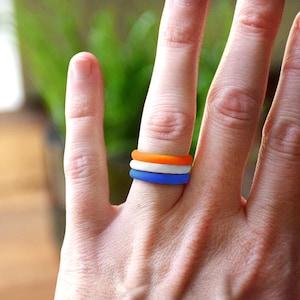 May include: Three silicone rings in orange, white, and blue are stacked on a finger. The rings are smooth and have a matte finish. The background is blurred, with green foliage visible.