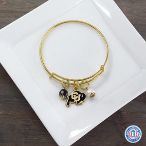 May include: Gold-tone bangle bracelet with three charms. The charms include a black and gold buffalo logo, a black and gold beaded charm, and a white pearl charm. The bracelet is displayed on a white square plate.