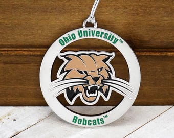 May include: A silver-colored Ohio University Bobcats ornament. The ornament features a circular design with the words "Ohio University" and "Bobcats" in green. The center showcases a brown and black bobcat.
