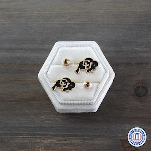 May include: A pair of gold-colored stud earrings with black enamel buffalo logos, displayed in a white velvet hexagonal jewelry box. The earrings are set against a dark wood grain background.