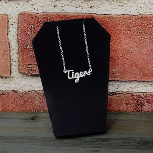 May include: A silver-colored necklace with the word "Tigers" in a cursive font. The necklace is displayed on a black jewelry stand against a brick wall background.