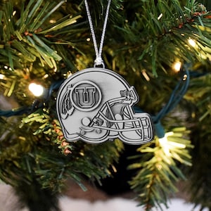 May include: Silver-colored Christmas ornament shaped like a football helmet. The helmet features a large letter "U" inside a circle, with a feather design on the side. The ornament hangs from a silver chain and is displayed on a Christmas tree.