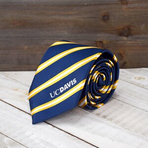 May include: Rolled navy blue tie with yellow and white stripes. The tie features the text "UCDAVIS" in white. The tie is made of a smooth fabric and is rolled up, showcasing the pattern and design.