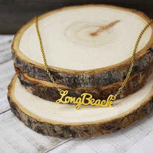 May include: A gold-colored necklace with the words "Long Beach" in a cursive font. The necklace is displayed on a stack of wooden slices with a natural, rustic appearance. The chain is delicate and the pendant is shiny.