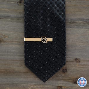 May include: A black tie with a repeating pattern, paired with a gold-colored tie clip. The tie clip features the University of Colorado Boulder logo in black and gold. The tie and clip are displayed on a wooden surface.